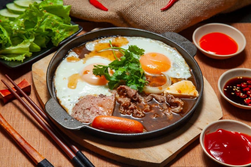 Chảo bánh mì với hai quả trứng ốp la, thịt bò, chả và xúc xích, bên cạnh là rau xà lách và mấy chén nước chấm